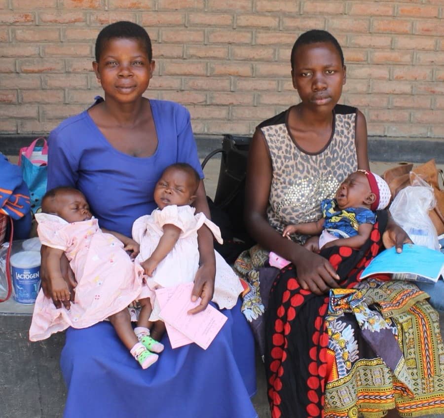 Two women sit against a brick wall with three babies: the left woman in blue holds two infants in pink outfits, the right woman in a patterned dress has a baby in blue on her lap.