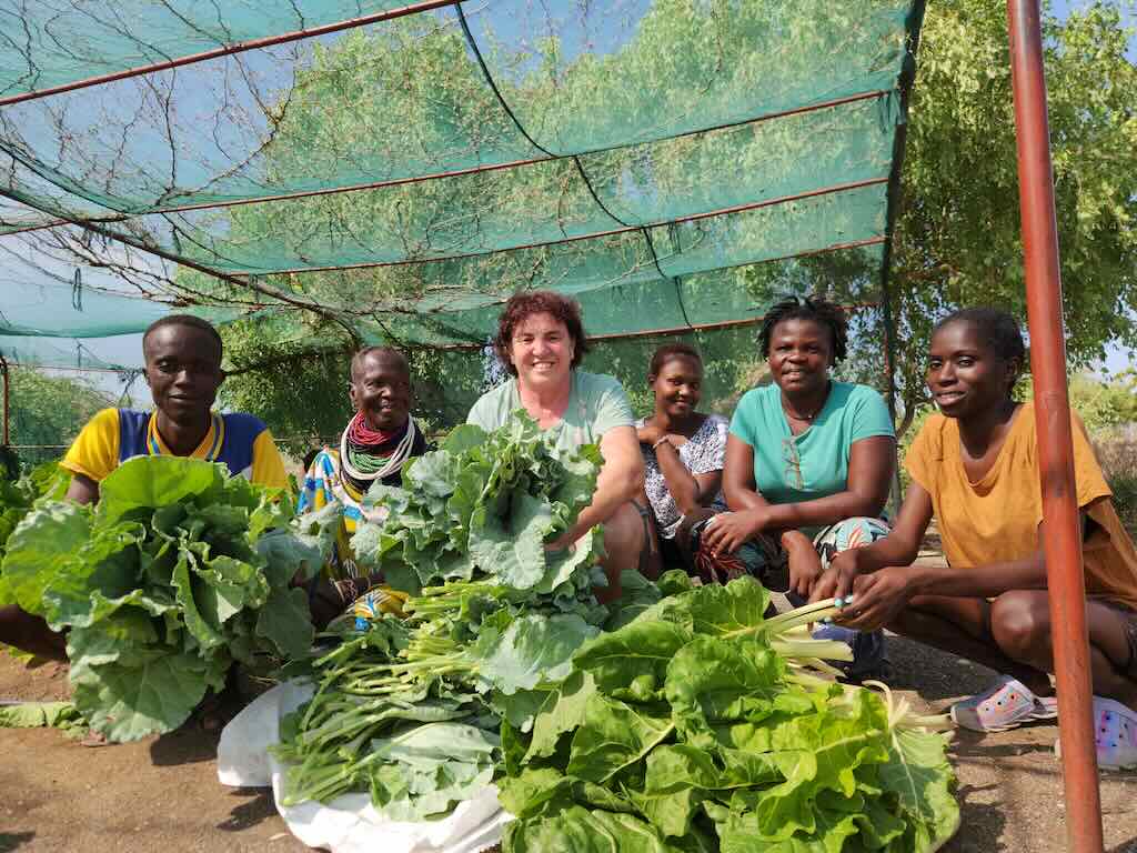 Group of people sitting under a green net canopy with large leafy greens gathered in front, posing with harvested vegetables in an outdoor garden.