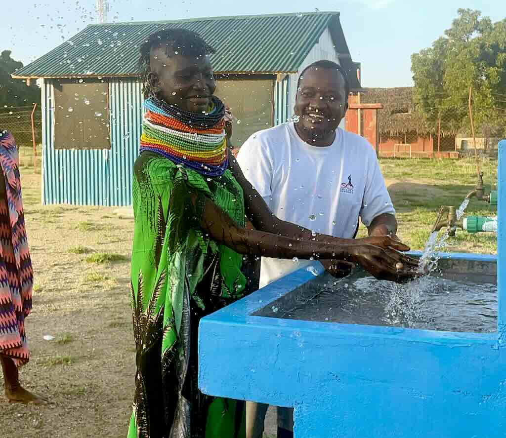 Two people stand at a blue water trough, smiling as water sprays from the tap.