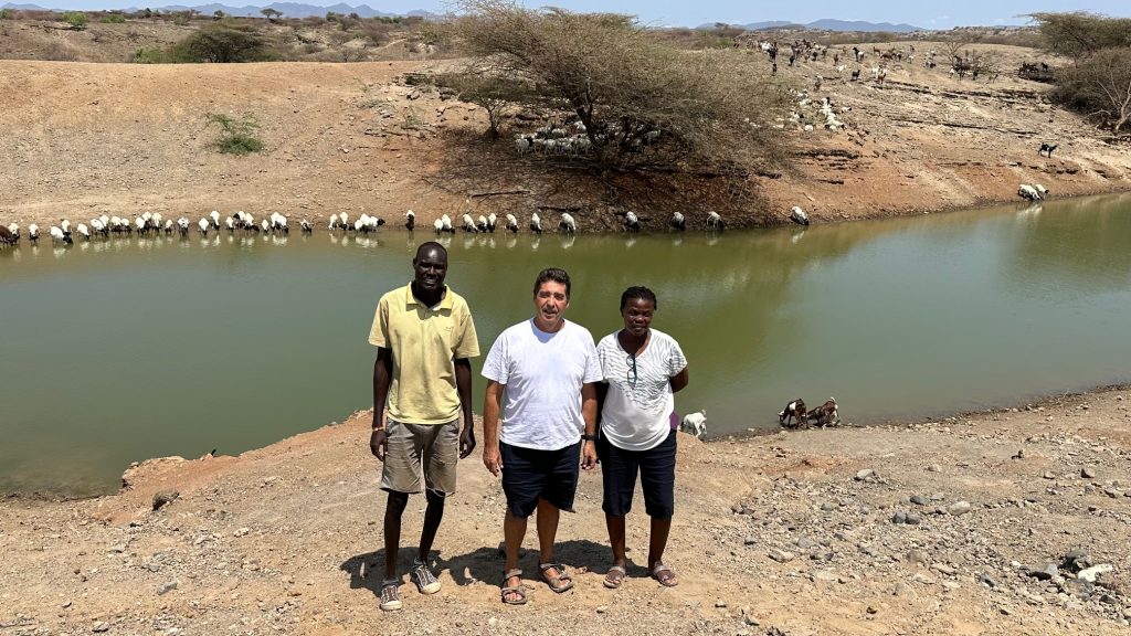 Three people stand near a green river bank, with a long line of sheep along the water and a dry, hilly landscape in the background.