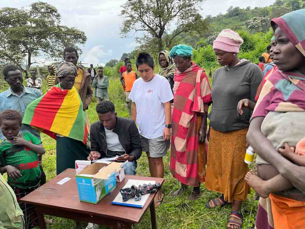 Man seated at a wooden table writes on paperwork while a crowd of villagers, many in bright wraps, stand around in a grassy field.
