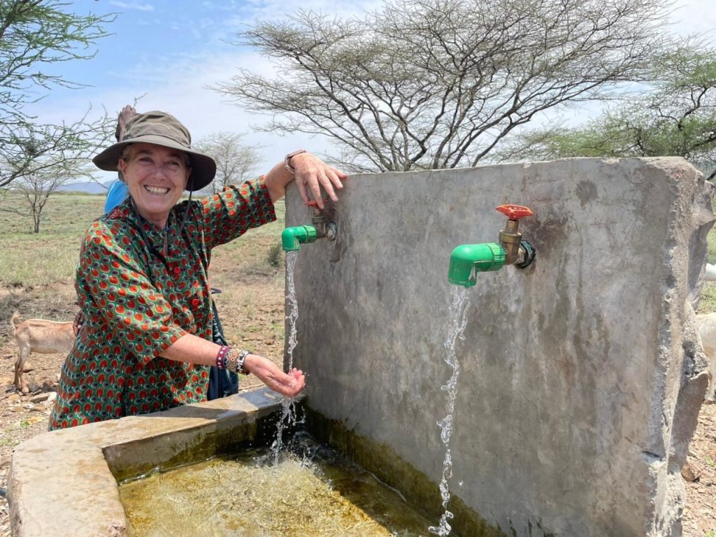 Smiling woman in a patterned dress and wide hat washing her hands at an outdoor concrete water trough with green taps.