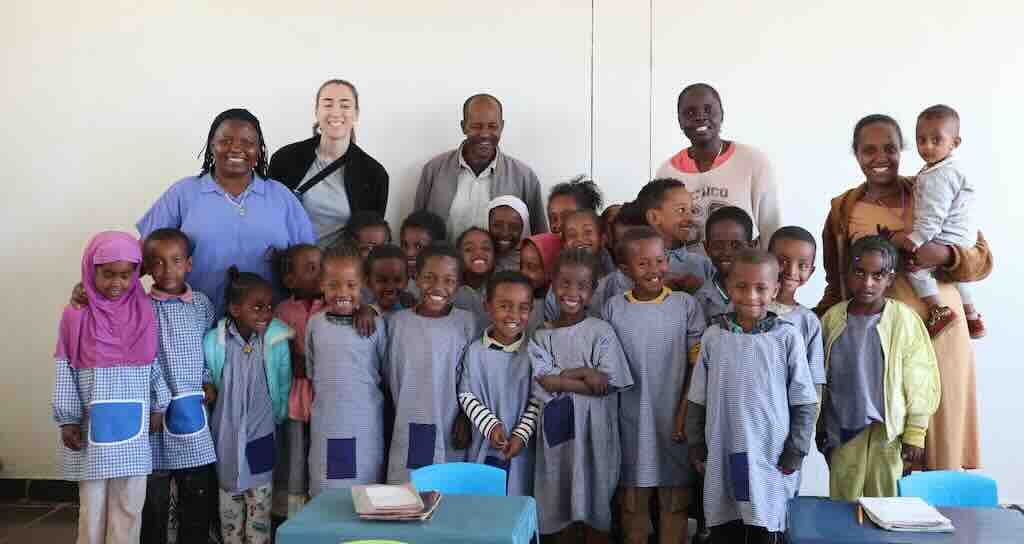 Group of smiling schoolchildren in blue uniforms posing with adults in a classroom setting, for a class photo.