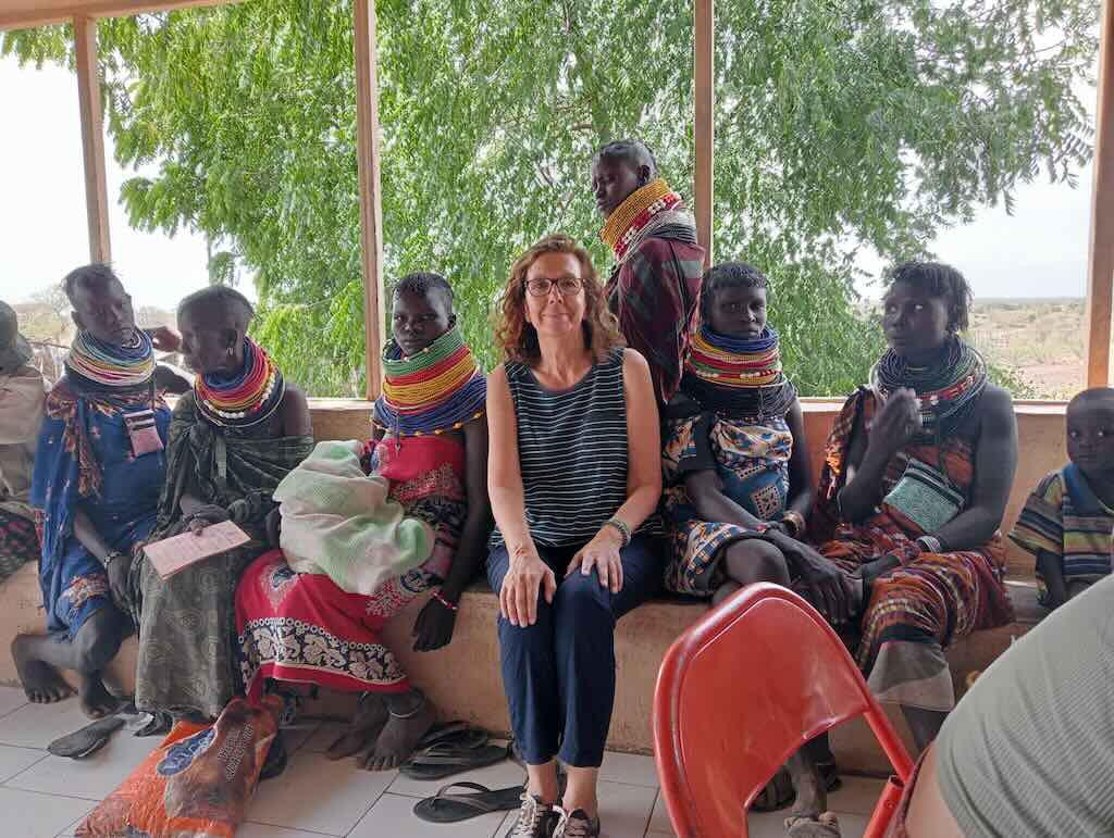 A light-skinned woman sits center, surrounded by Maasai people wearing colorful bead necklaces and wraps in an open shelter.