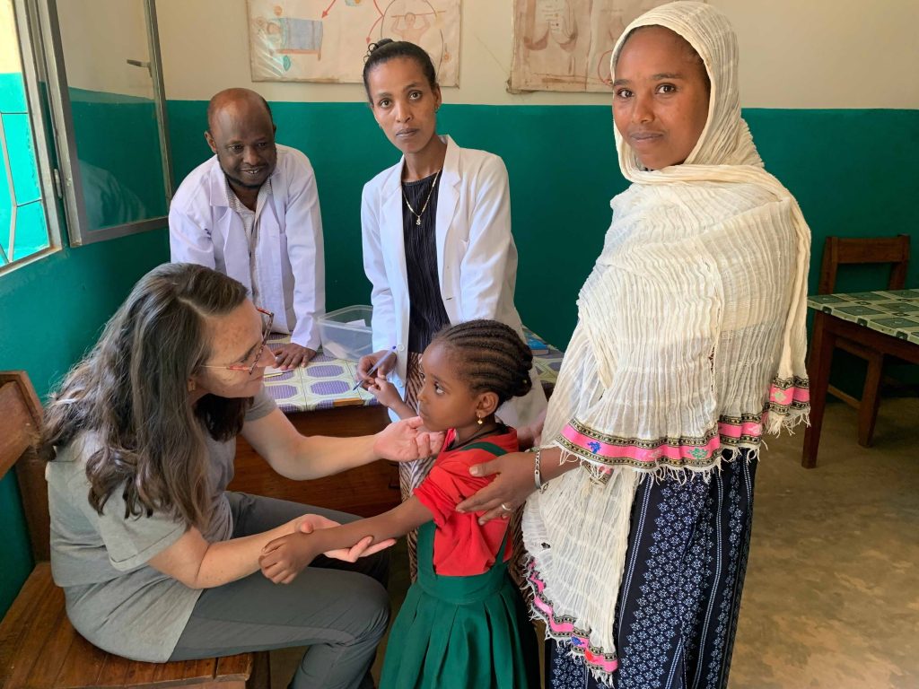 A nurse seated at a table checks a child’s arm while two health workers and a woman in a light scarf look on in a clinic room.