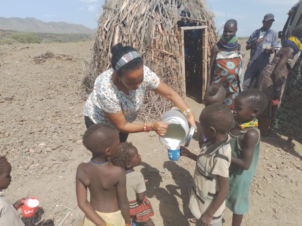 Woman pours milk from a metal pitcher into a blue cup for a line of children in a dry, rural village setting with a thatched hut nearby.