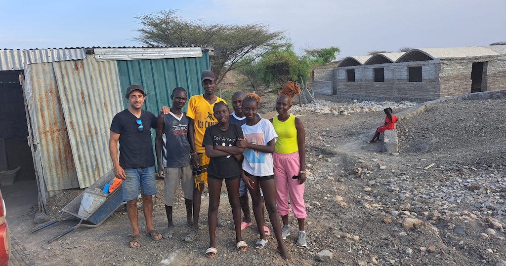 A diverse group of seven people standing together on a rocky construction site with unfinished brick buildings in the background.