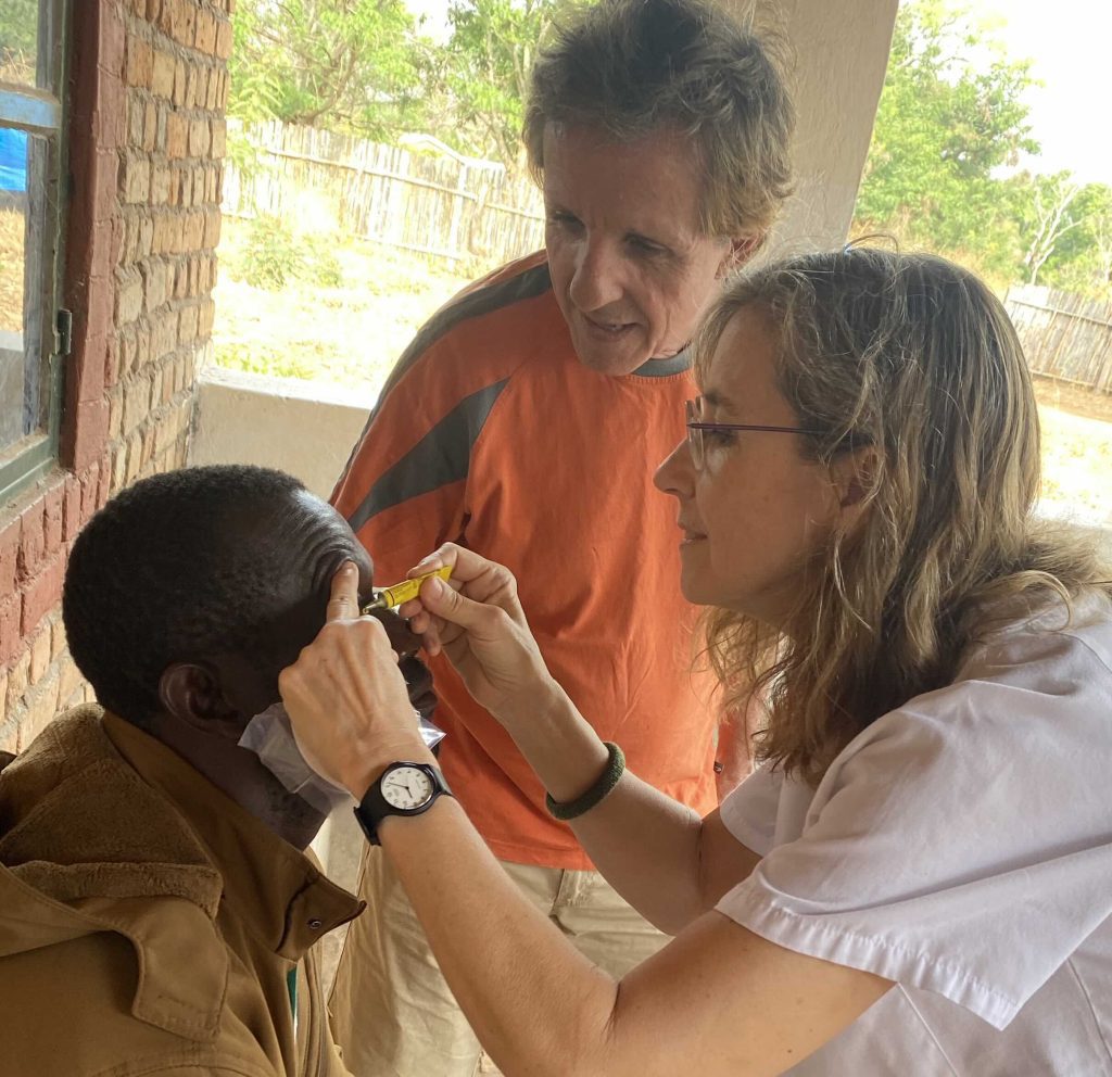 Three people—an adult man in an orange shirt, another adult wearing glasses, and a child—gather closely as they work with a yellow tool near the child’s head on a shaded porch, with a brick wall to the left and greenery in the background.