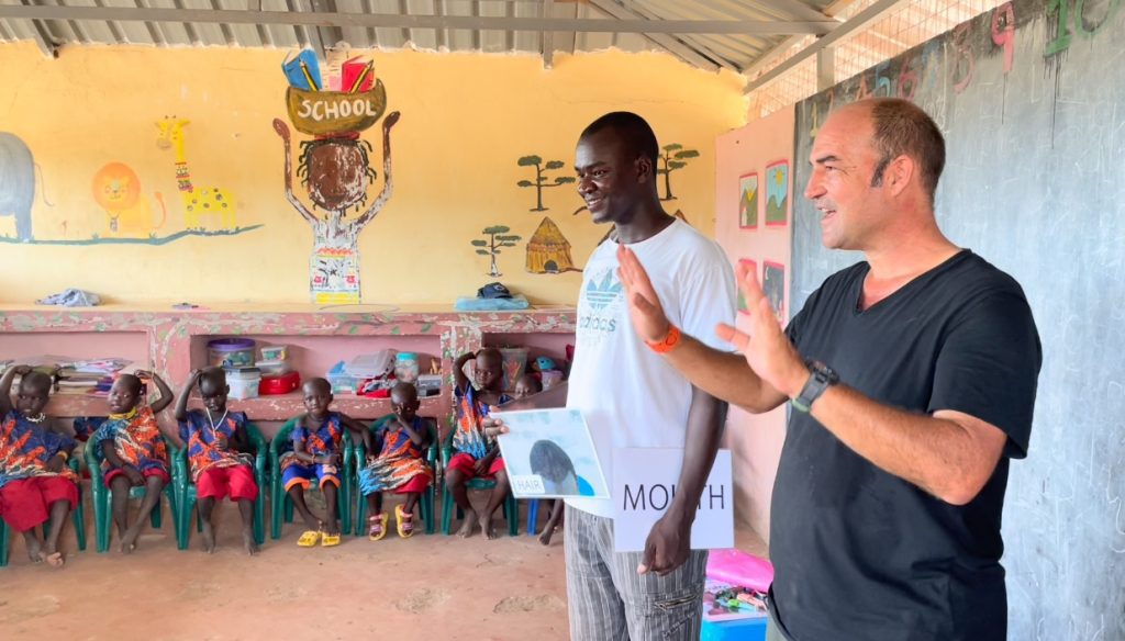 Two men stand at the front of a classroom as young children sit in green chairs, one holding a card labeled HAIR and the other a sign reading MOUTH during a body-parts lesson.