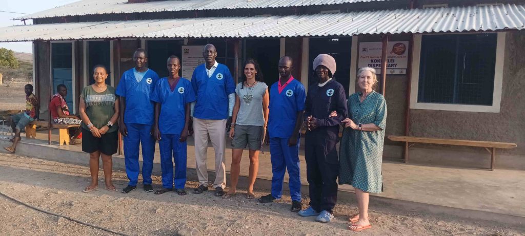 Group of people standing in a row outside a building, some in blue medical scrubs, smiling at the camera.