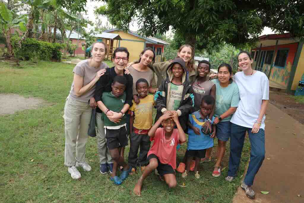 Group of volunteers and local children posing on a grassy schoolyard with colorful huts in the background.