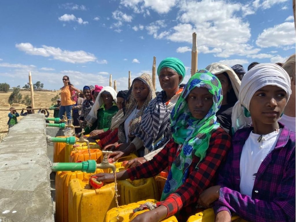 Group of women and girls queueing at water taps outdoors, filling yellow containers in a dry, sunny landscape.
