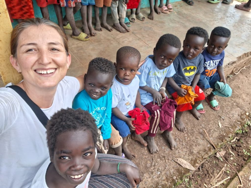 Smiling woman taking a selfie with a row of barefoot children sitting on the ground, holding colorful clothes outdoors.