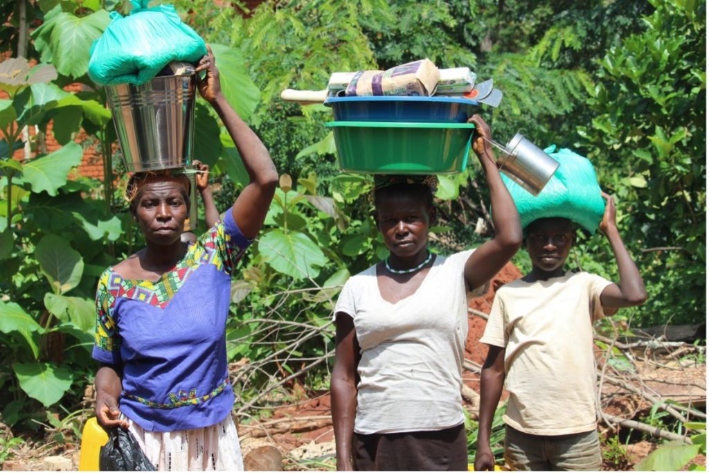 Three people walk in a garden carrying bowls and bags on their heads, balancing supplies outdoors.