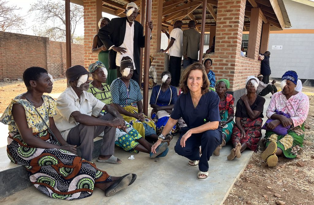 Healthcare worker in navy scrubs kneels with a community of people with facial bandages outside a brick building, showing support and care.