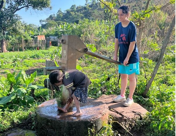 Two children in a lush garden operate a hand water pump; a girl stands on a platform turning the lever while a boy puddles water at the water basin.