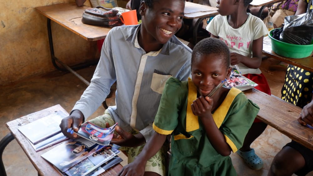Smiling man shows a photo booklet to a young student in a green and yellow school uniform in a classroom.