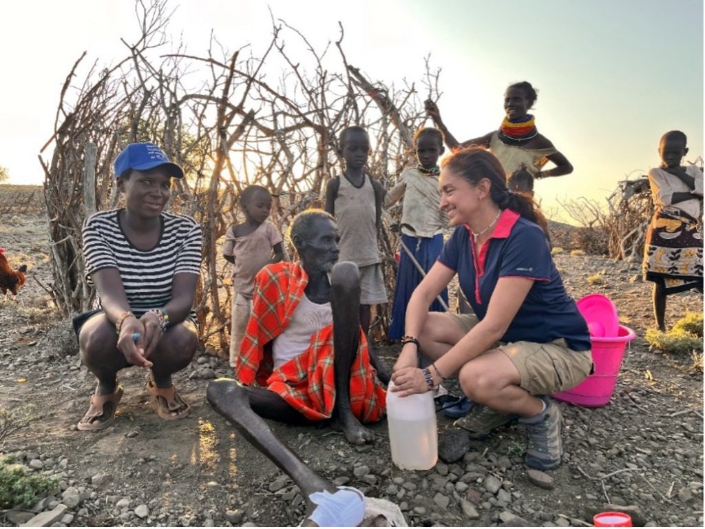 Volunteer woman kneels beside an elderly man, pouring water from a bottle in a dry rural setting as children watch nearby.