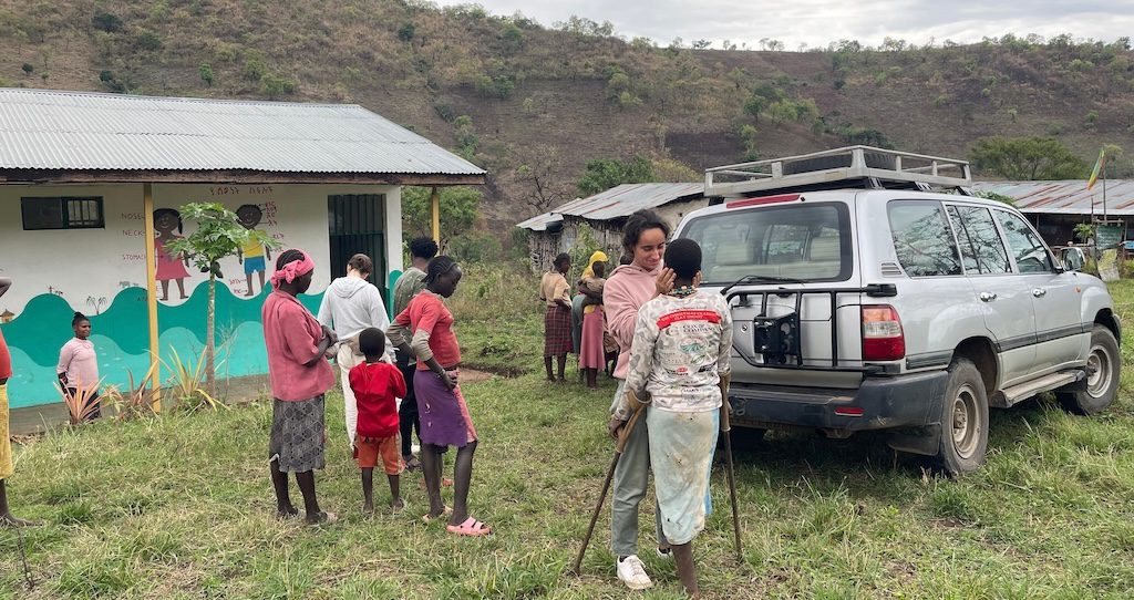 A group of people, including children, gather outside a small clinic with a mural, while a silver SUV with gear is parked nearby in a grassy area under cloudy skies.
