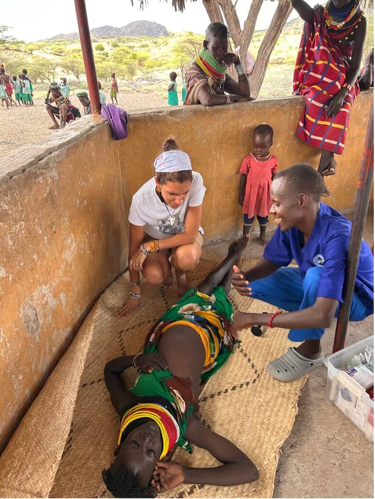 Volunteer kneels beside a person lying on a woven mat as a caregiver in blue tends to them in a dusty village courtyard with onlookers nearby.