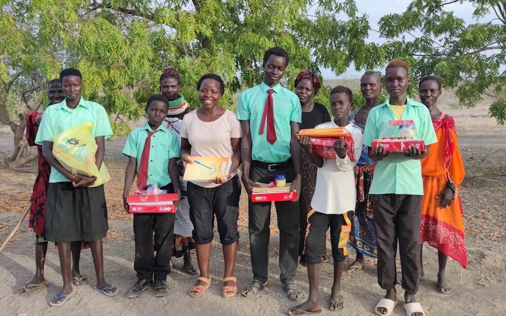 Group of students standing outdoors under a leafy tree, each holding school supplies and boxes of materials for learning.