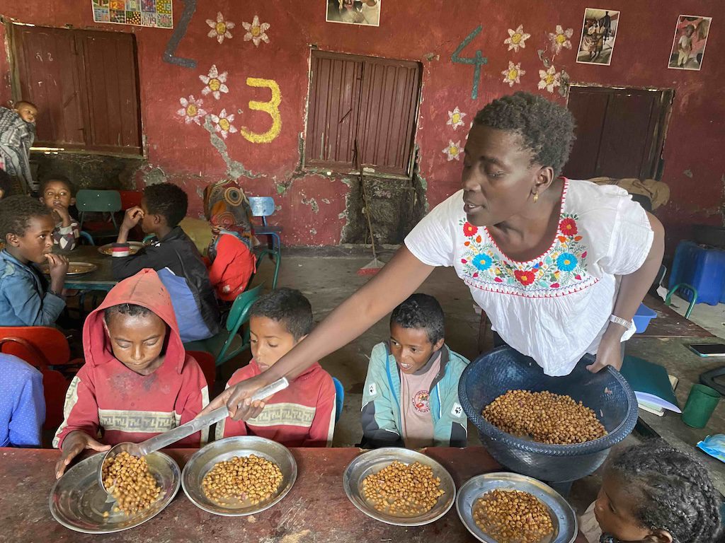 Teacher serves bowls of beans to children at a table in a classroom setting with colorful walls and numbers behind her.