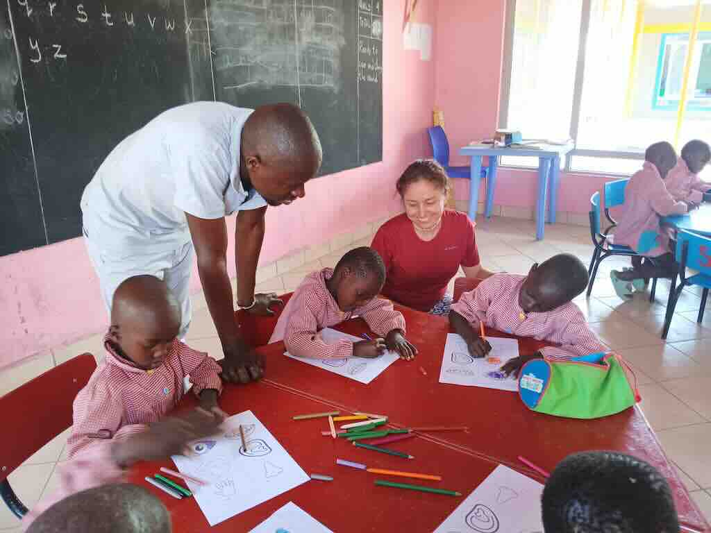 Teacher helps several young students color worksheets at a red table in a bright pink classroom, with pencils scattered between them.