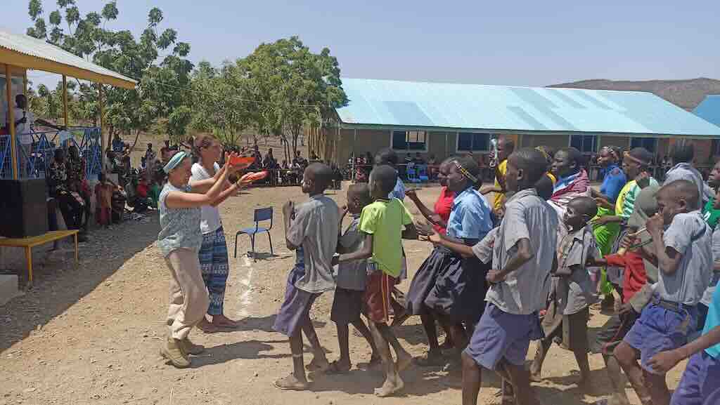 Adults lead a lively dance with a crowd of schoolchildren in a dusty outdoor yard near blue-roofed buildings.
