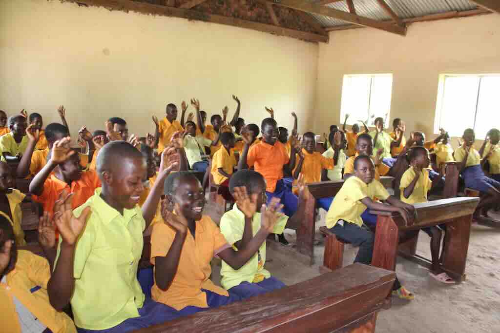 Group of schoolchildren in a simple classroom, raising hands while seated on wooden benches.