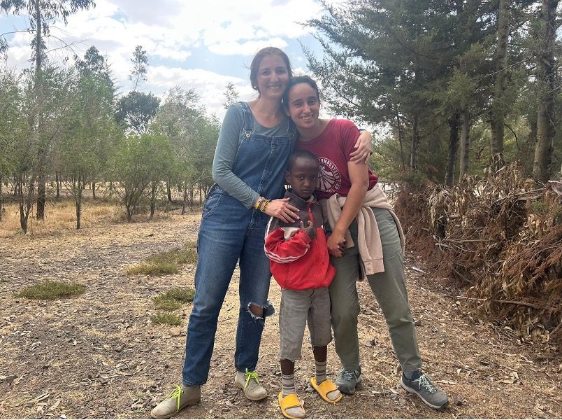 Two women and a young boy pose together outdoors, smiling and hugging on a dirt path among trees and brush.