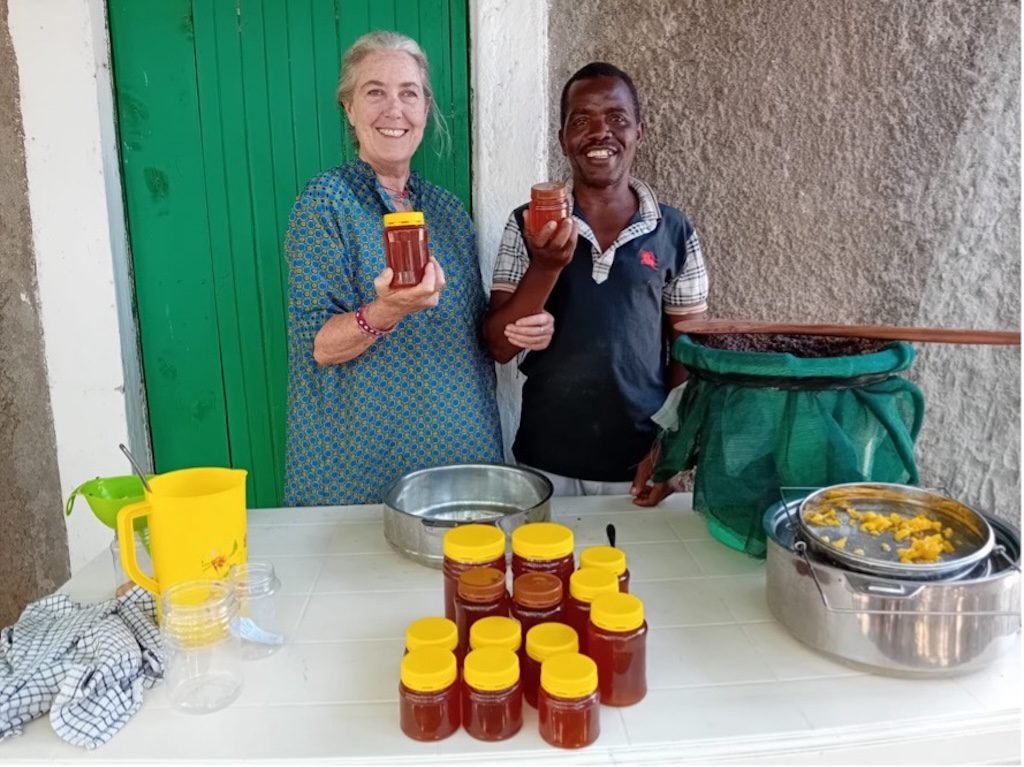 Two smiling vendors stand behind a table of honey jars, displaying small glass jars with yellow lids outdoors next to a metal pot and a green bag-lined barrel setup.