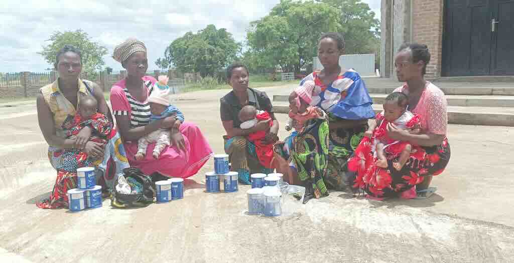 Mothers with babies sitting on a street, with blue cans of infant formula arranged in front of them.