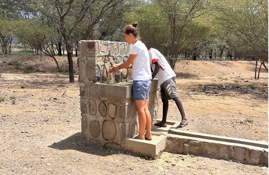 Two people refill water at a stone outdoor drinking fountain in a dry, sandy area; one woman in a white shirt and blue shorts is using the tap.