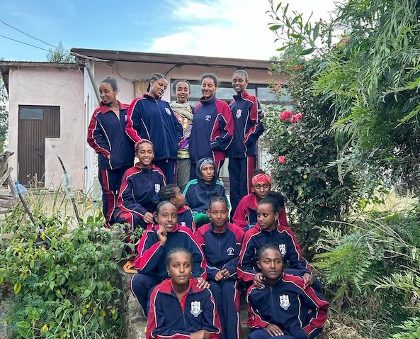 Group of students in matching navy and red tracksuits posing outdoors on a garden path in front of a small building.