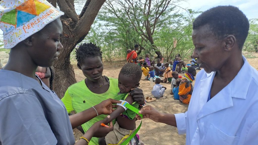 Two health workers measure a child's arm with a tape while a mother holds the toddler, outdoors under a tree.