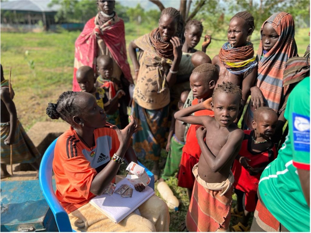Young woman in an orange Adidas shirt sits on a blue chair distributing or counting money and papers to a crowd of children and adults nearby in a rural setting.
