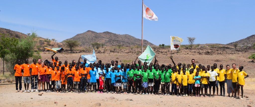 Group of children in orange, blue, green, and yellow shirts posing in a dry, rural landscape with hills and flags behind them.