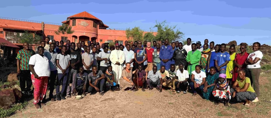 Large group of men and women posing outdoors in front of a red-orange building on a sunny day, a rural setting behind them.