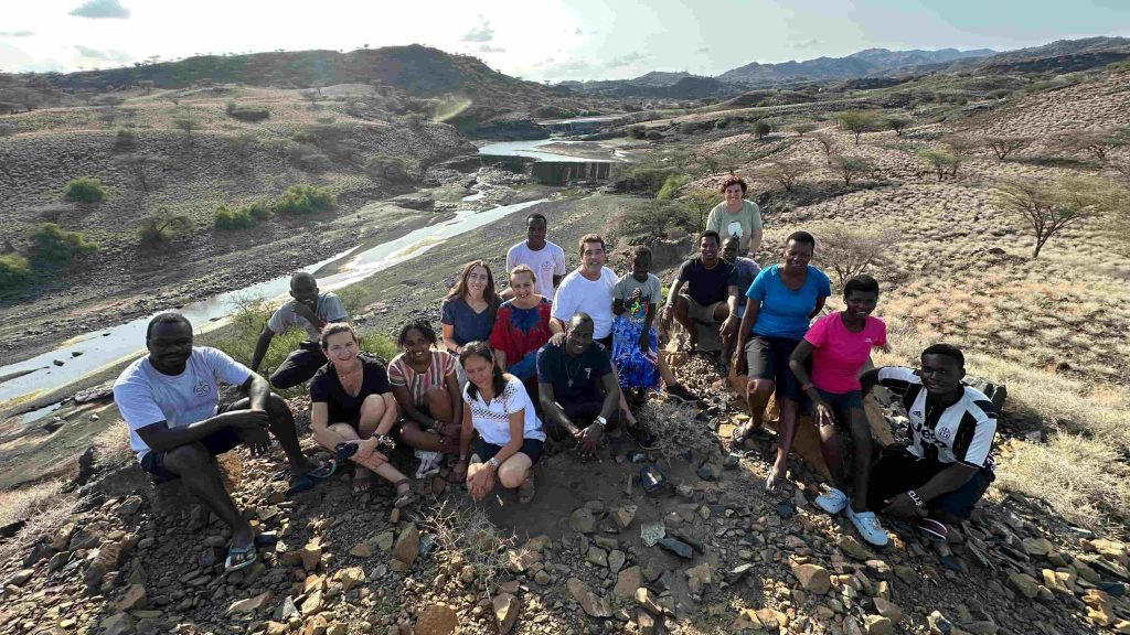 Group of about 15 people posing on a rocky hillside with a river valley and distant hills in the background.