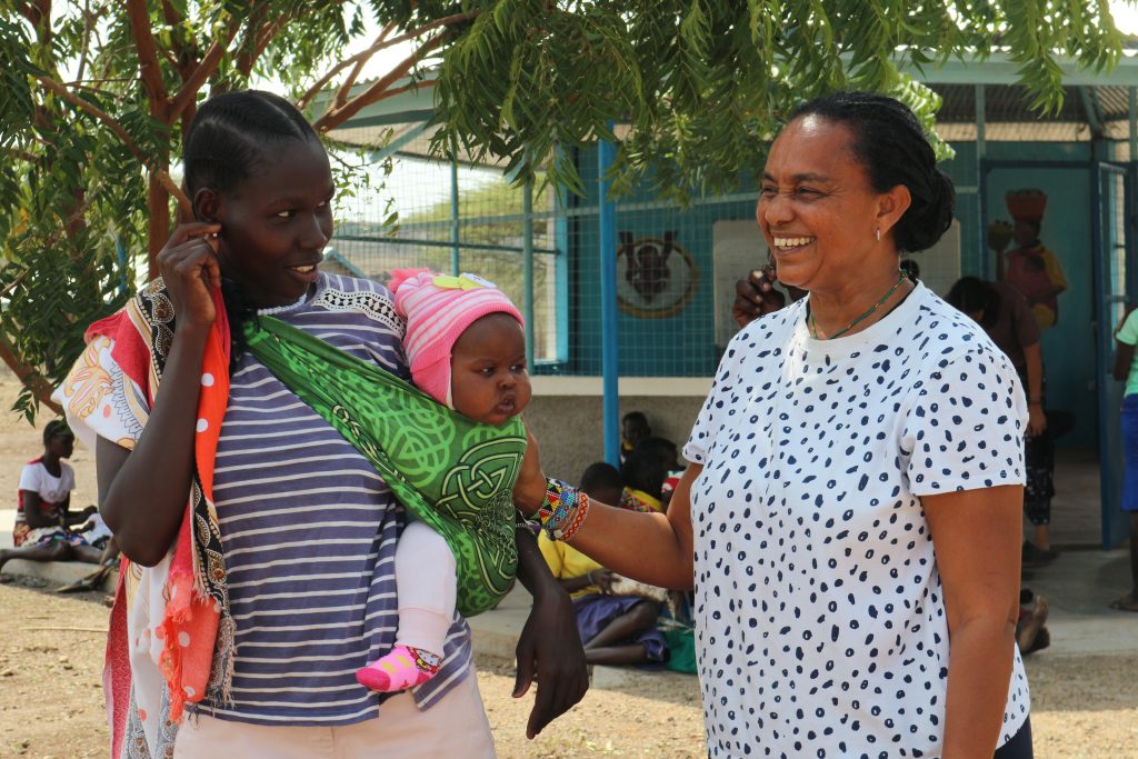 Two women converse outdoors as one carries a baby in a green patterned sling between them, smiling under a tree.