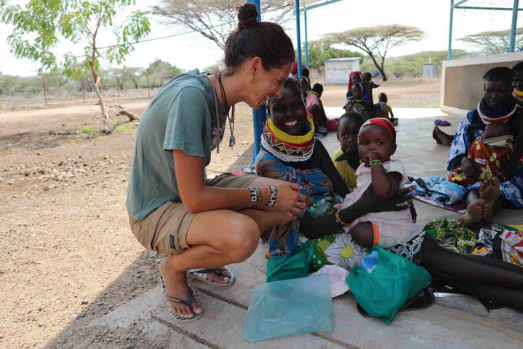 Woman crouching and smiling at local Maasai women and children wearing colorful beadwork outdoors, during a communal gathering.