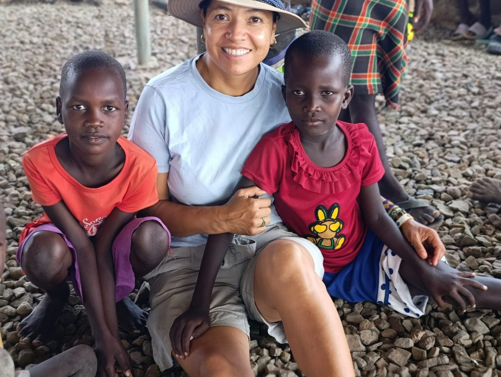 Smiling woman wearing a hat sits between two children on a rocky outdoor surface, all facing the camera.