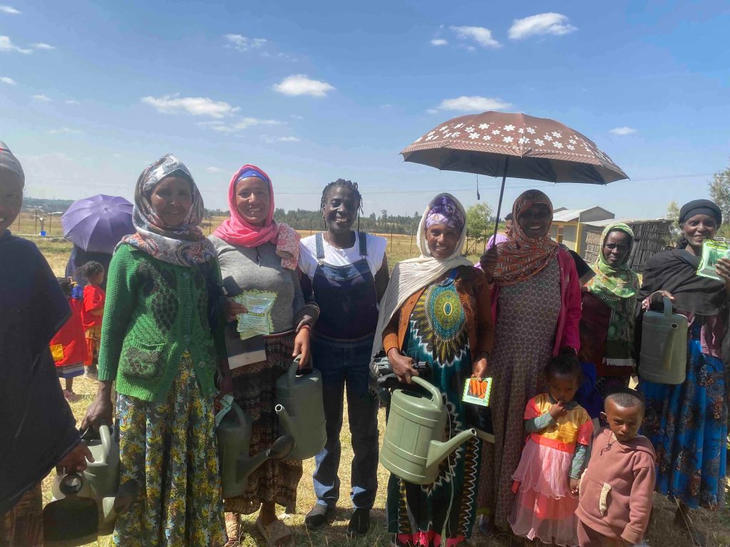 Group of smiling people outdoors, women and children in colorful clothing, some holding green watering cans, under a bright blue sky with a small umbrella visible.