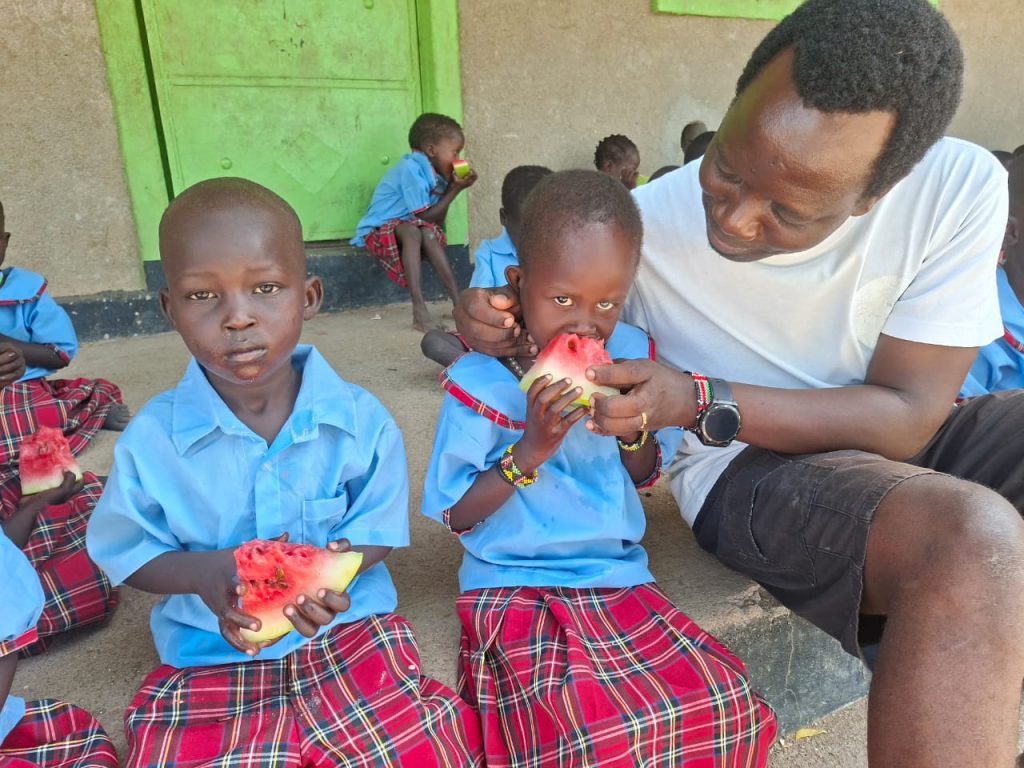 A man in a light shirt sits with young children in blue uniforms eating watermelon, outdoors at a school setting.