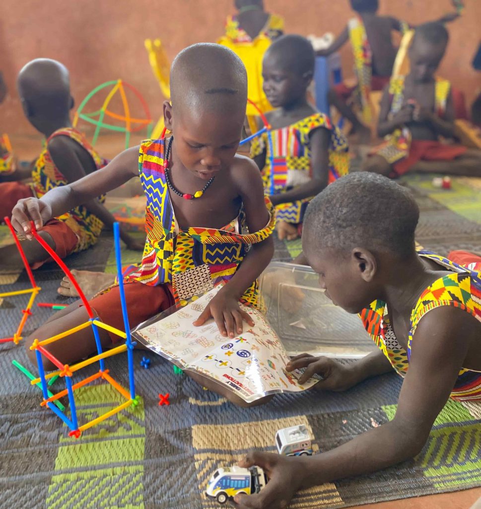 Children in bright, patterned clothes build with colorful plastic construction sticks on a woven mat indoors, with drawings and toys nearby.