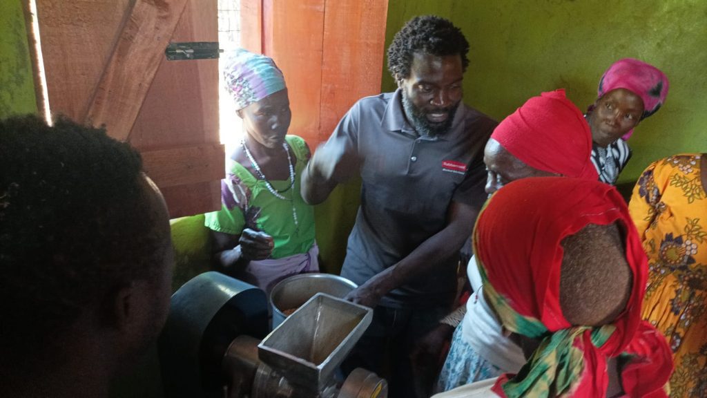 A man in a gray polo explains a metal grinding/milling machine to a group of women in colorful headscarves inside a green-walled room.