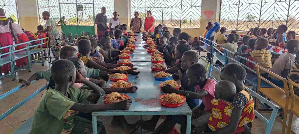 Children sitting at long tables in a bright cafeteria, eating from red bowls during a communal meal at a relief center or schoolhouse.
