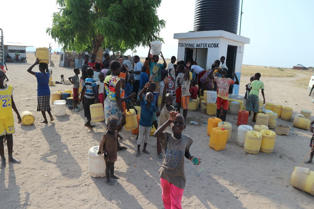 Group of children around a water kiosk with many plastic jerrycans; a large black water tank sits on a white building labeled 'Todonyang Water Kiosk' in a dusty, rural area.