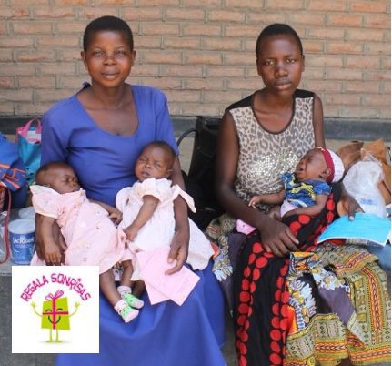 Two women sit against a brick wall holding four infants; one wears blue, the other a patterned dress, with a charity logo in the bottom-left.