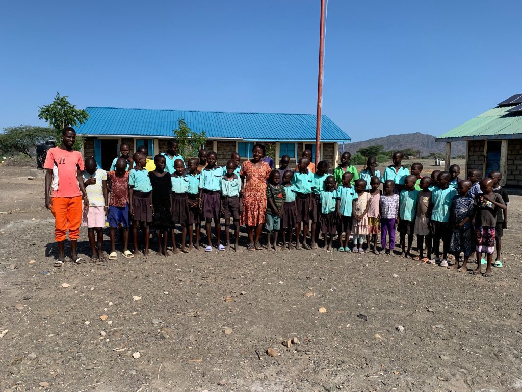Group of schoolchildren and a few adults standing in a line outside a building with a bright blue metal roof, posing for a photo.
